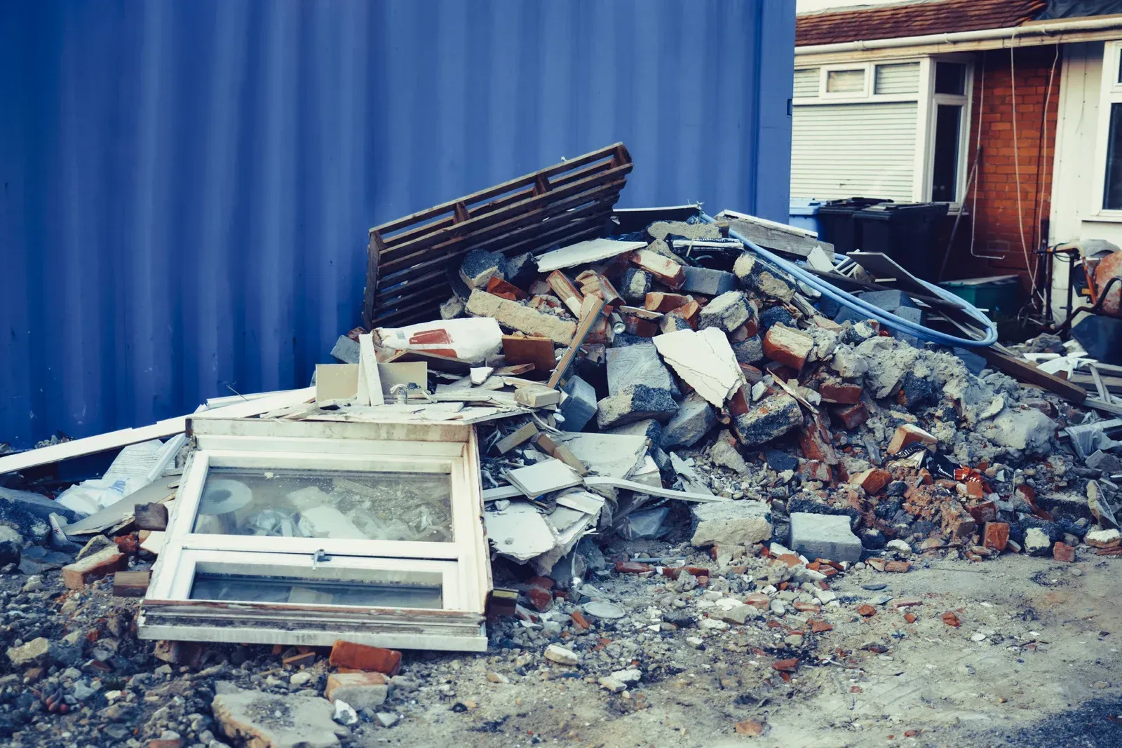 A pile of construction debris including broken bricks, rubble, and a discarded white window frame near a blue wall.