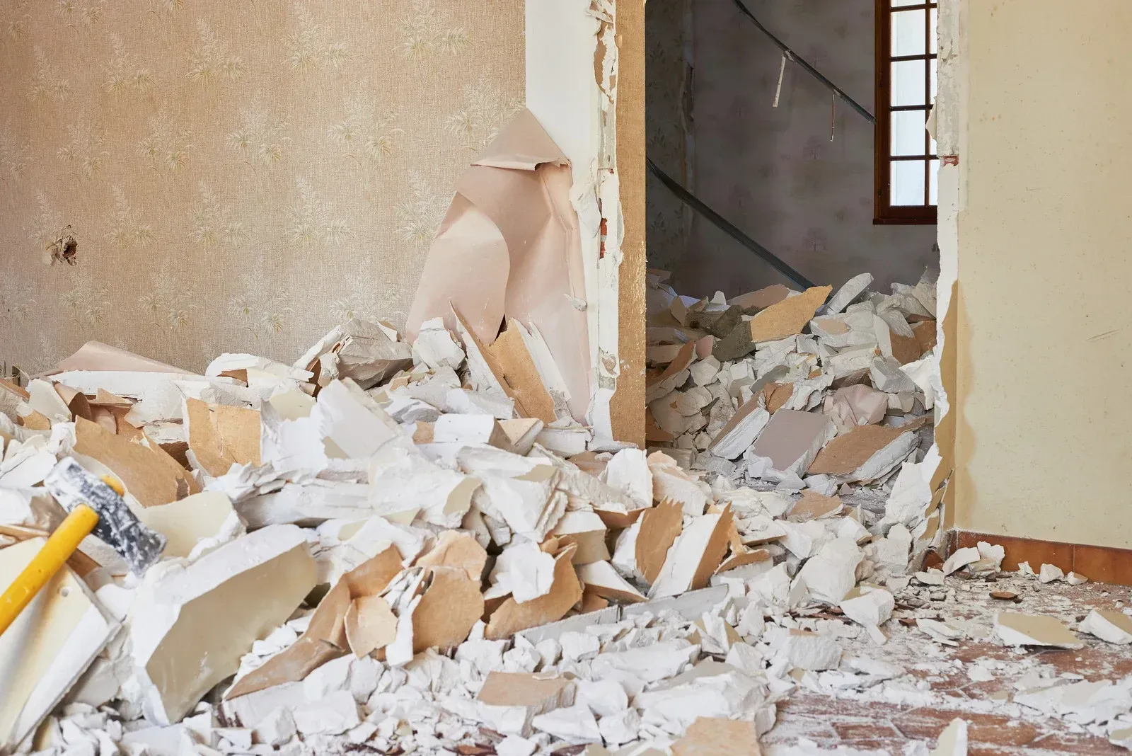 A room interior under renovation, featuring a pile of broken drywall debris on the floor and a hammer near a doorway.