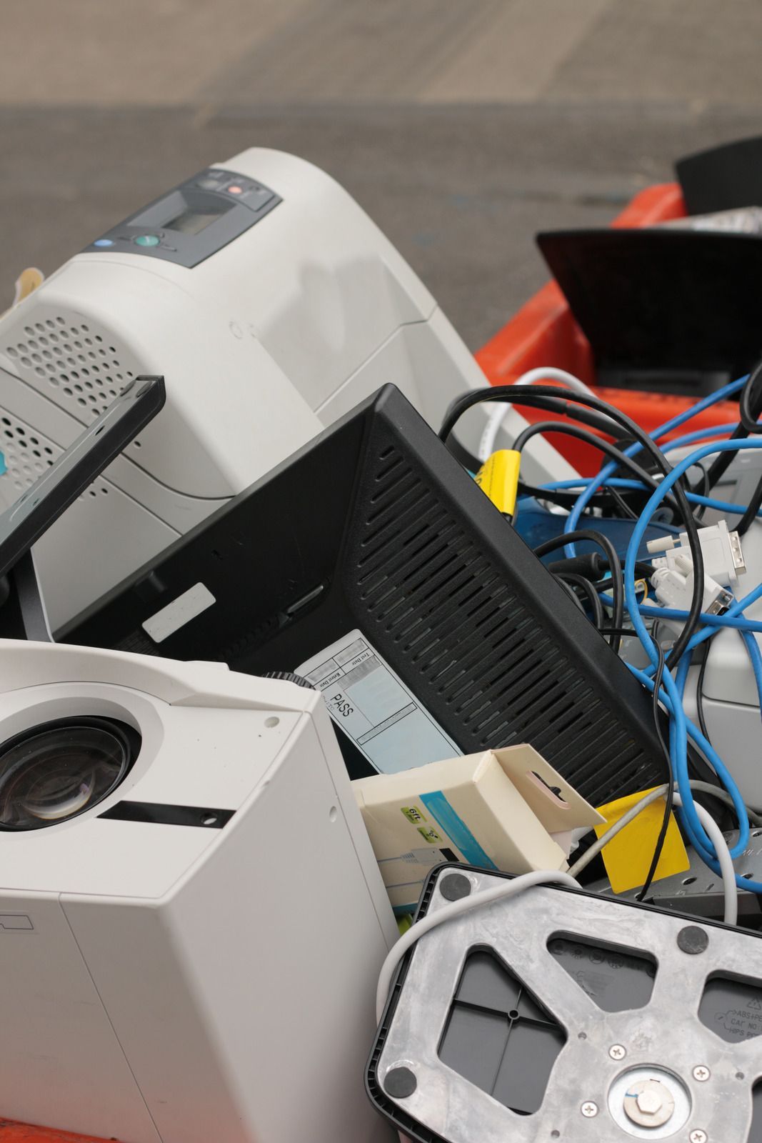 A pile of discarded electronic waste, including cables, a monitor, and printers in an orange bin.