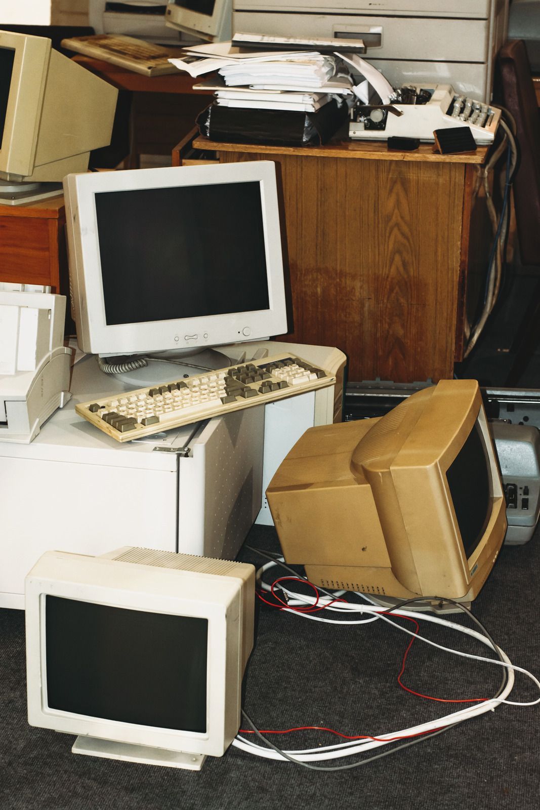 Several vintage beige computer monitors, a keyboard, and messy paperwork cluttered on a desk and floor.