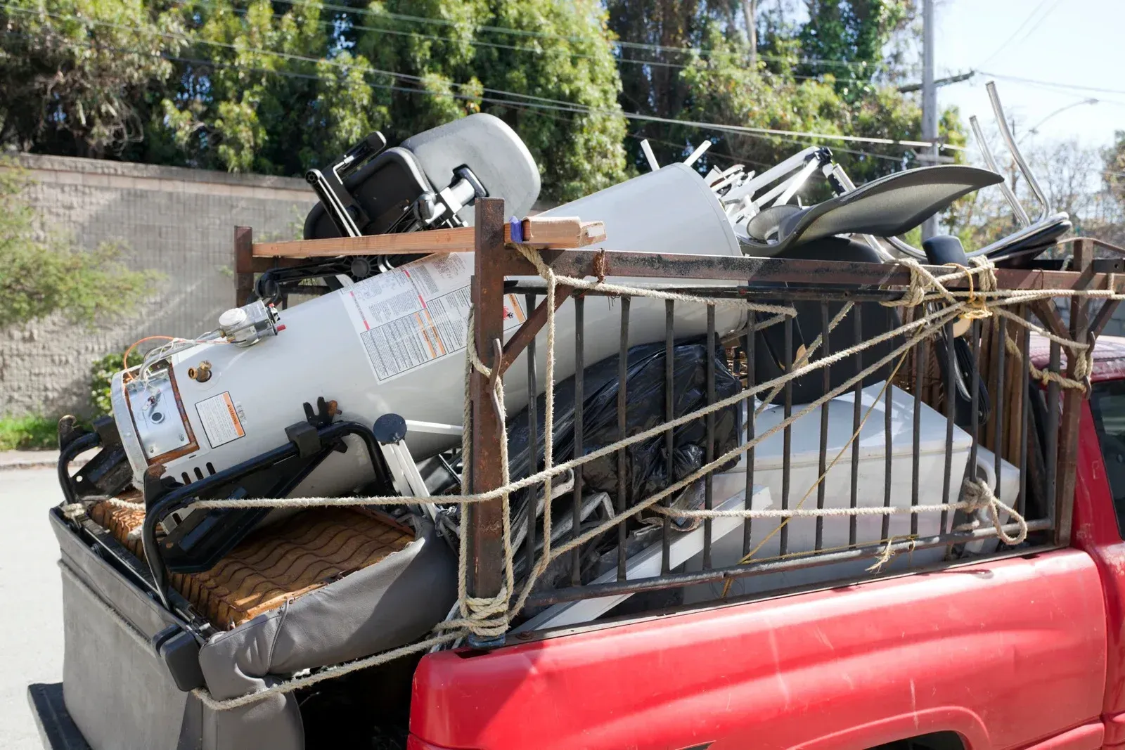A red pickup truck bed is heavily loaded with a white water heater, metal framing, and various pieces of discarded junk.