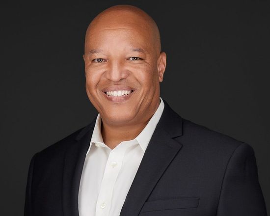 Man in black suit and open-collar white shirt, smiling against a dark gray background.