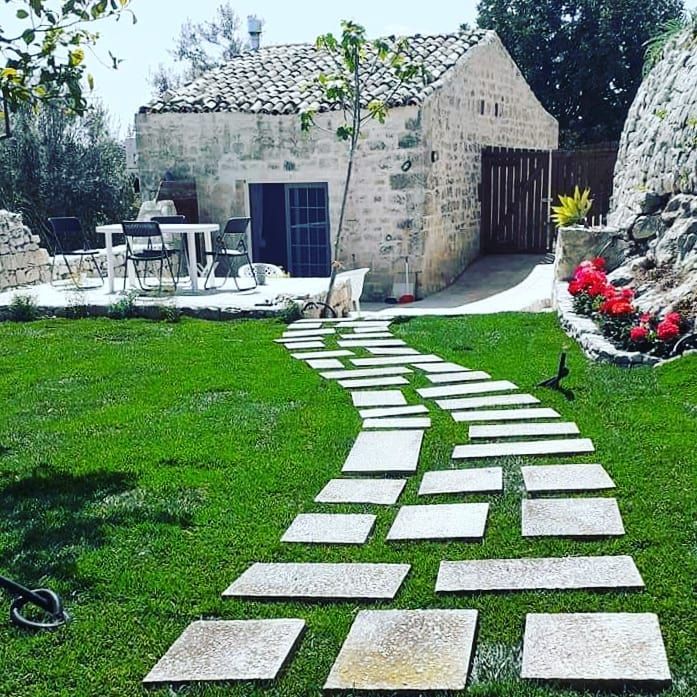 Sentiero in pietra che conduce a una rustica casa in pietra con prato, posti a sedere all'aperto e fiori rossi.