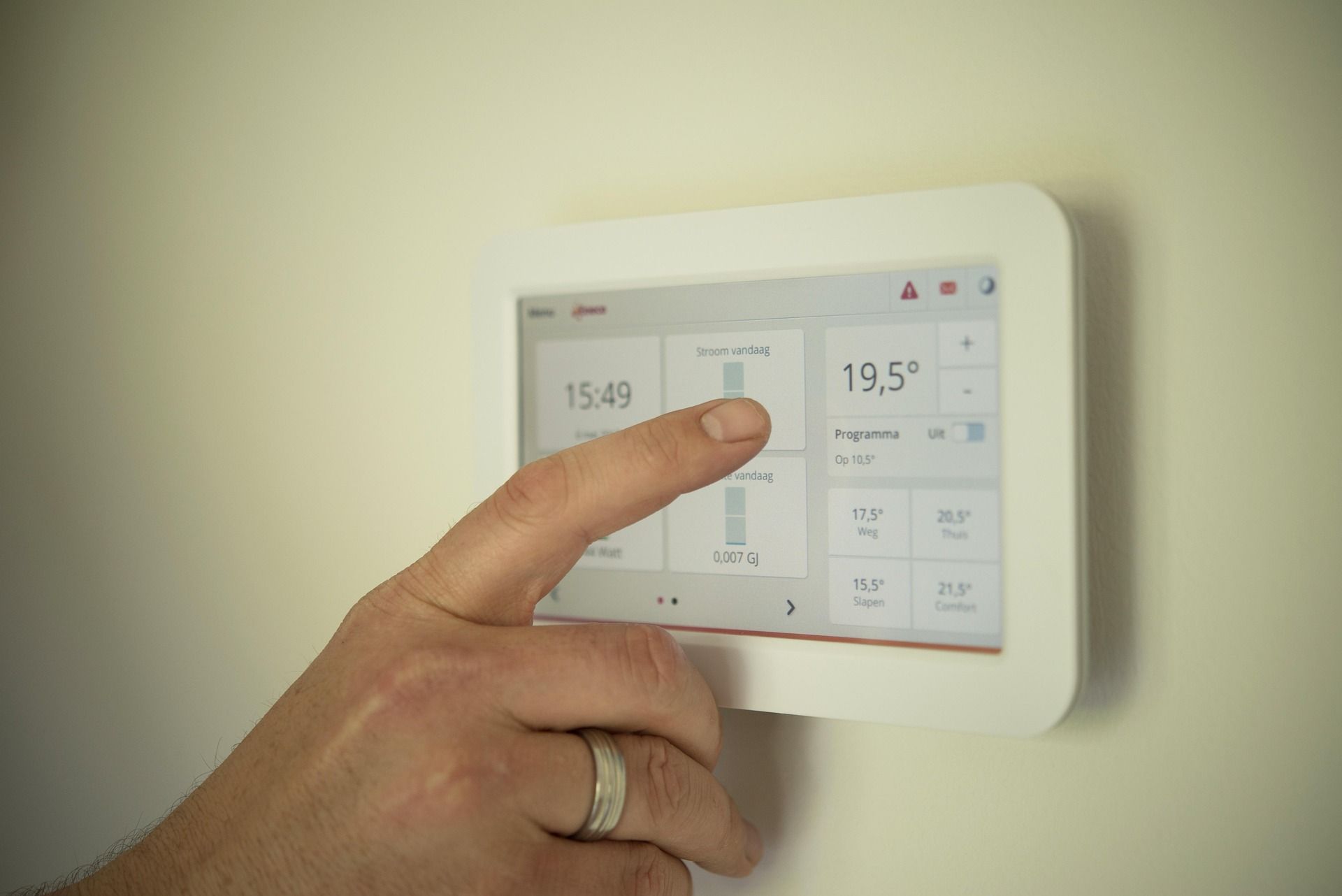 A hand interacts with a wall-mounted smart thermostat display showing a temperature of 19.5 degrees and the time 15:49.