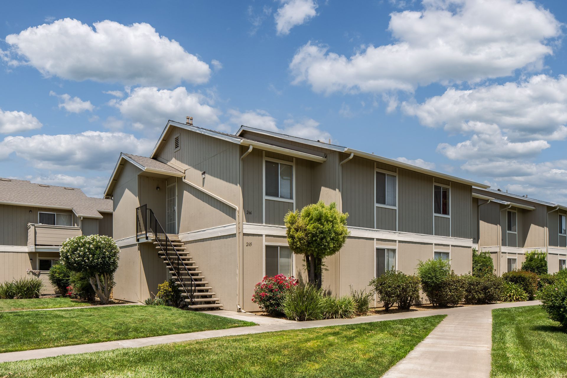 Two-story beige apartment building with green lawn, concrete path, and blue sky with clouds.