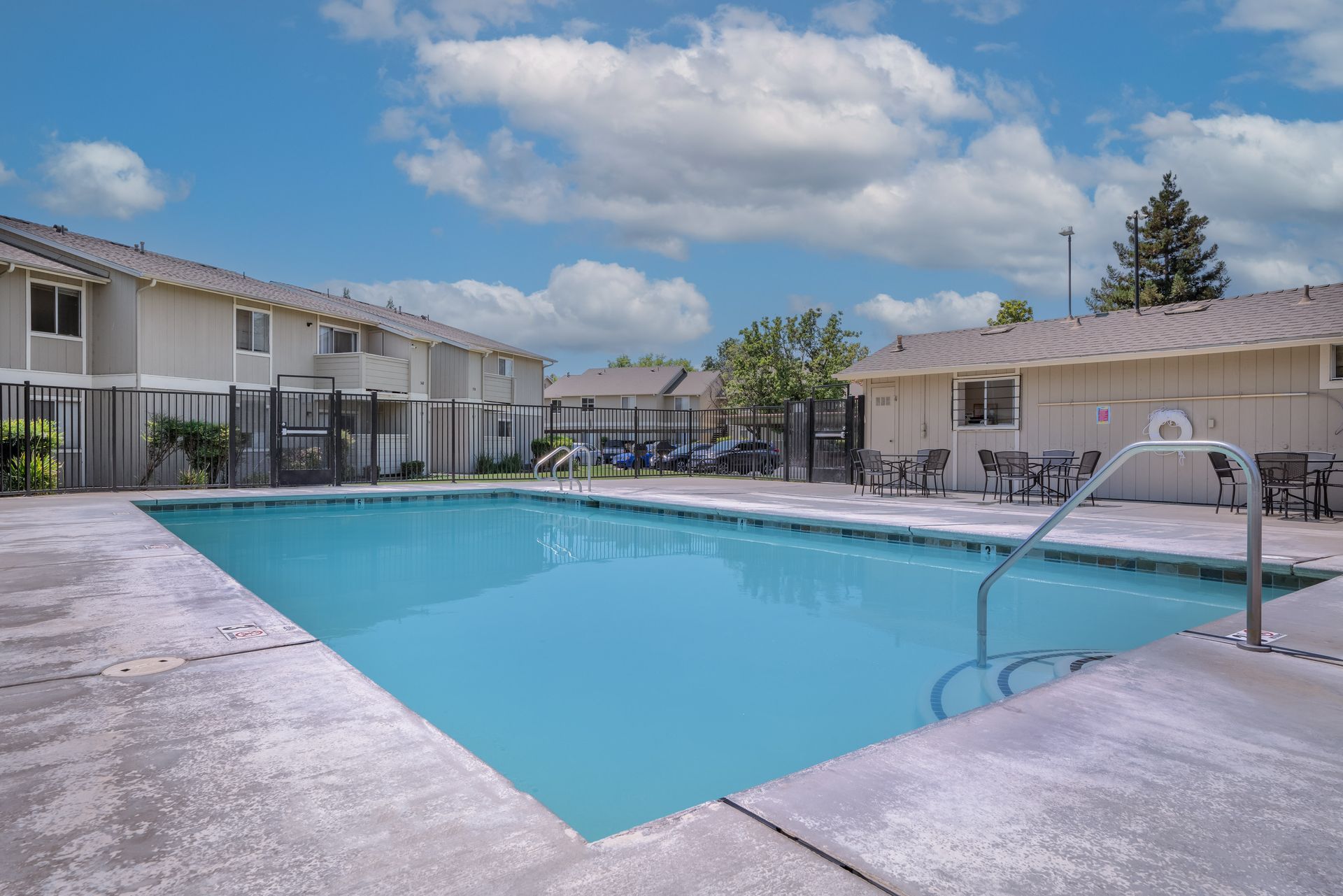 Swimming pool with lounge chairs and two-story apartment building in the background under a cloudy sky.