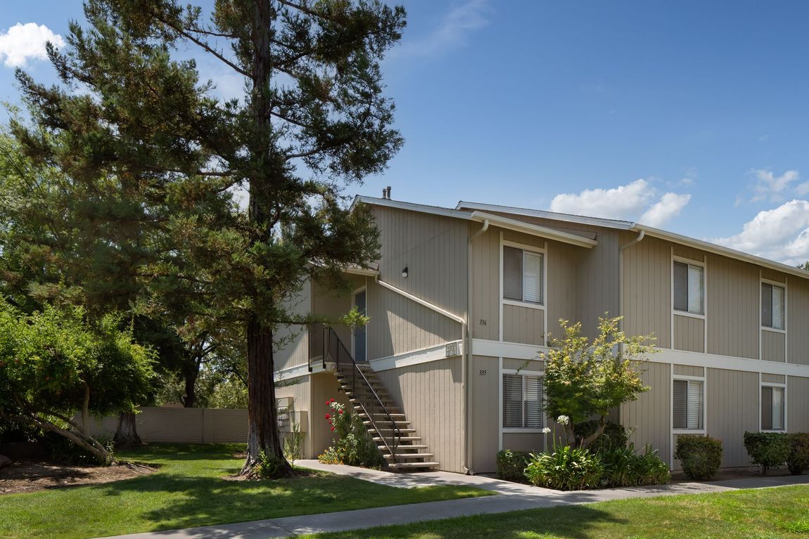 Two-story beige apartment building with outdoor staircase and trees under a blue sky.