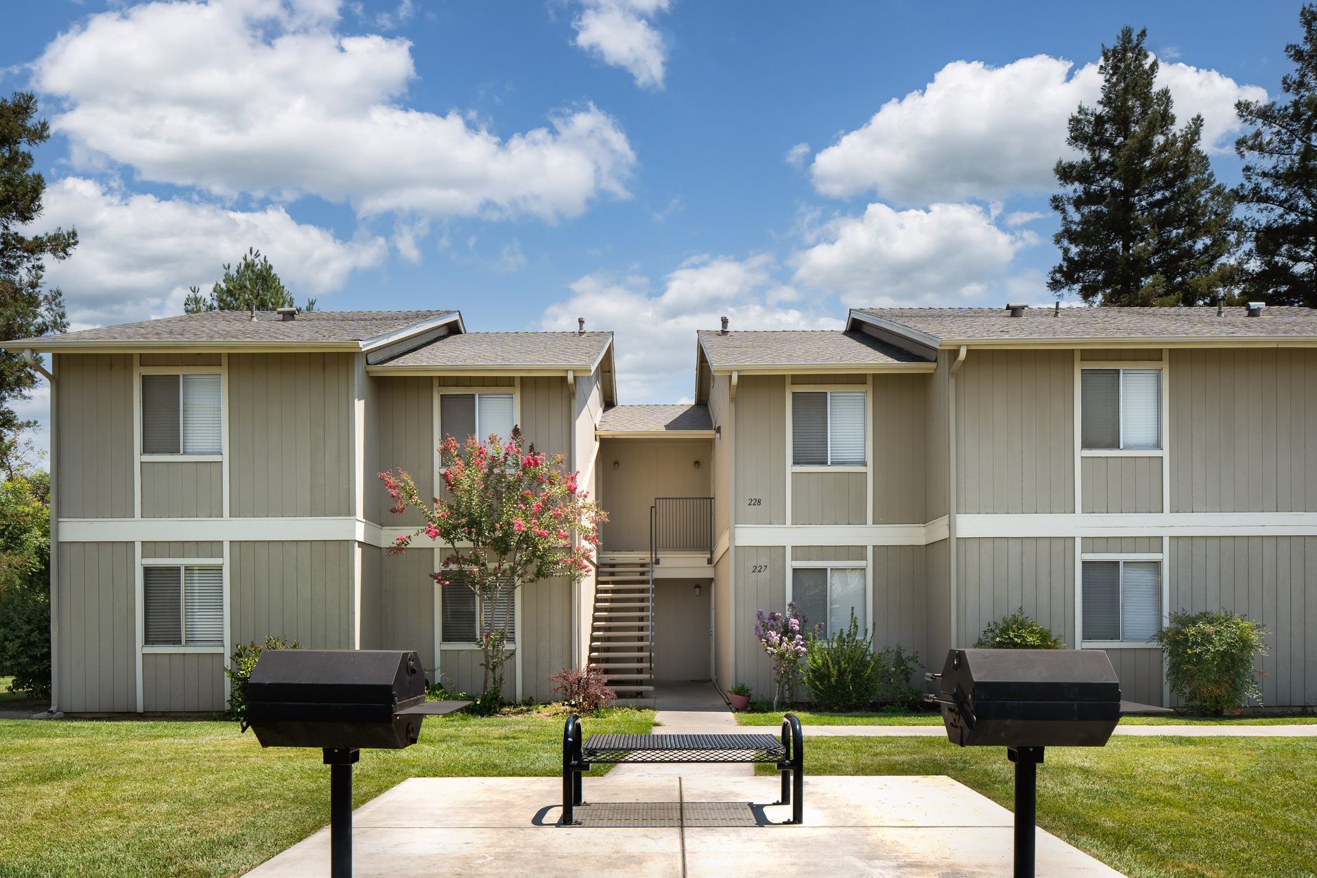 Apartment complex exterior with grills, bench, and blue sky.