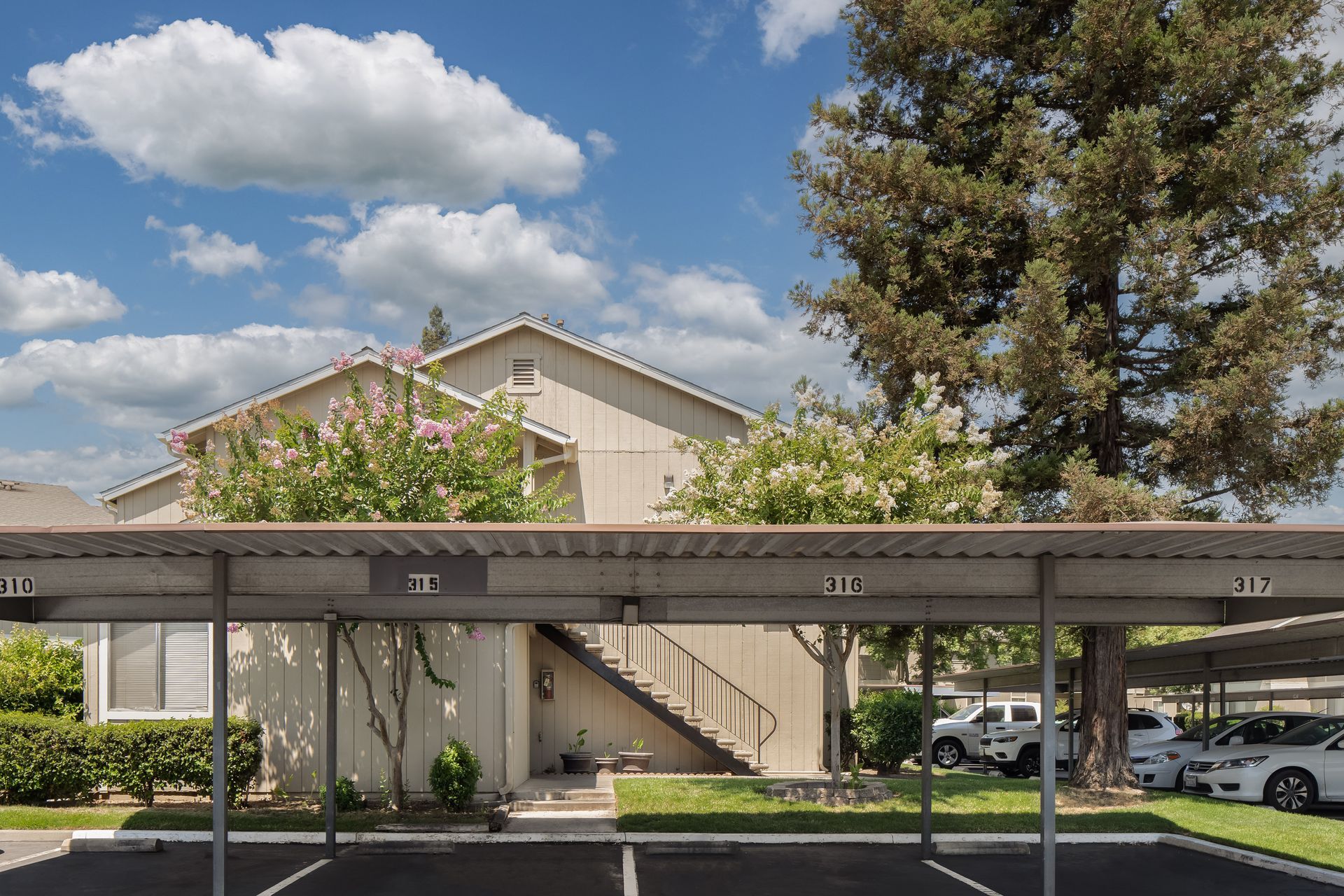 Apartment complex with parking, covered by carports. Tan building, blue sky, trees.