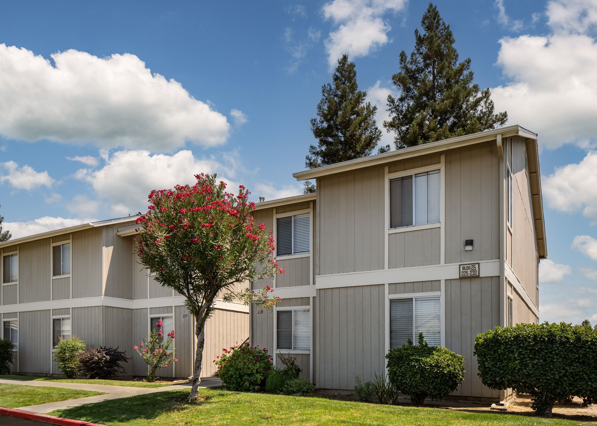 Two-story gray apartment buildings with white trim under a blue sky. Green grass and bushes in front.