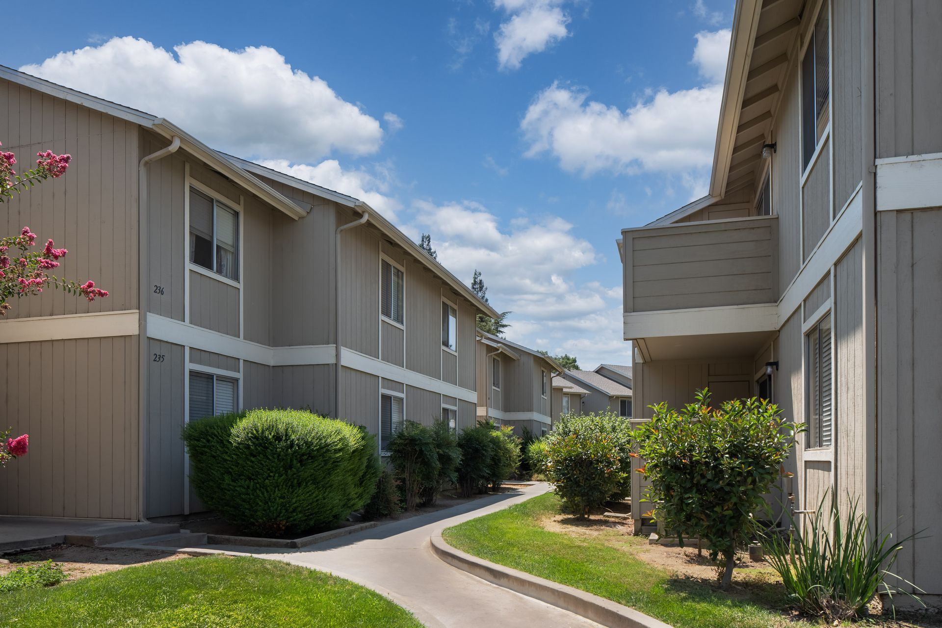 Apartment complex with beige buildings, green bushes, and a walkway under a blue sky with clouds.