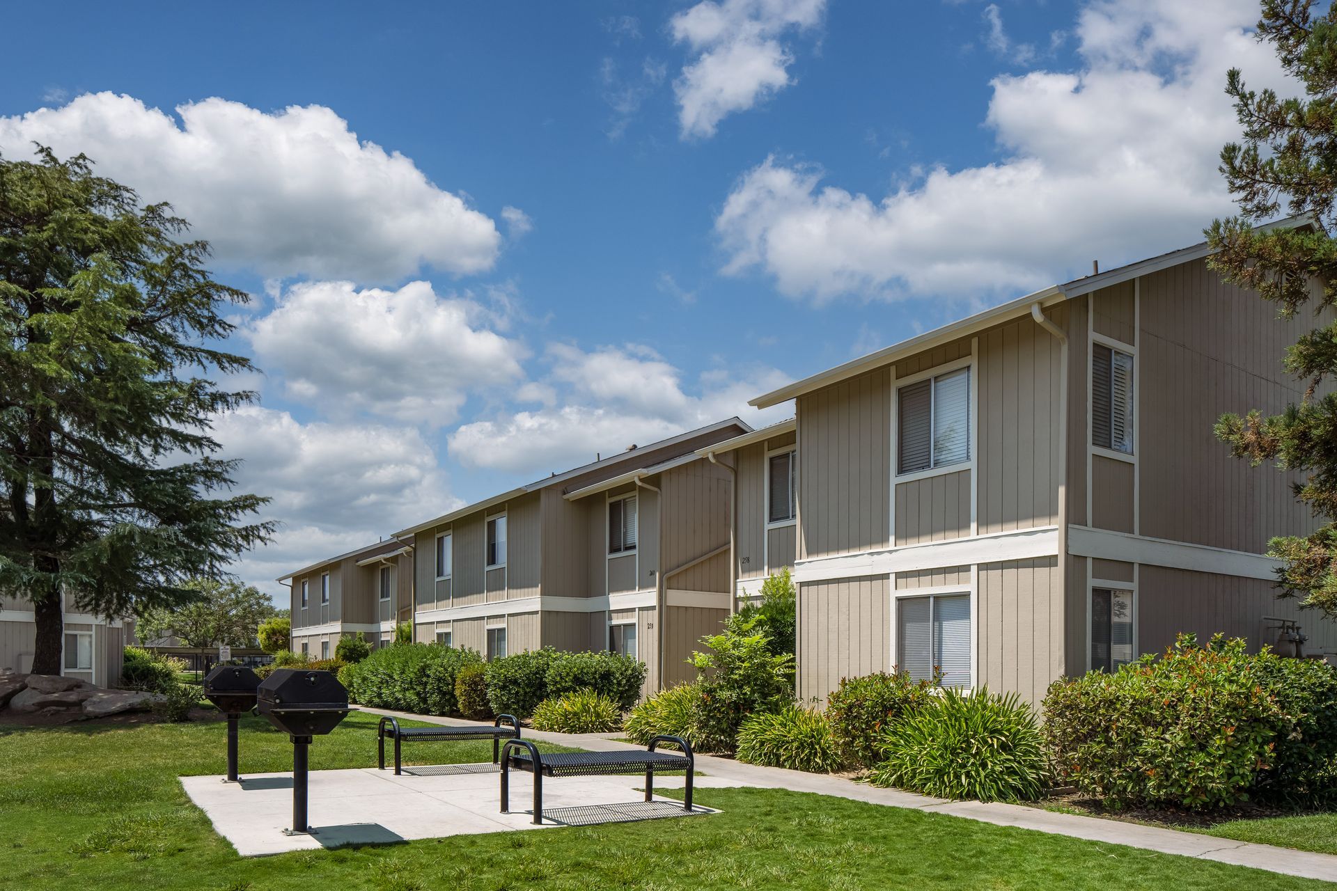 Two-story apartment buildings under a bright blue sky with barbecue grills in the foreground.