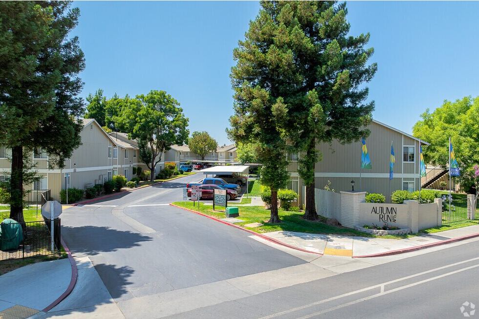 Apartment complex entrance on a sunny day. Buildings are light grey, surrounded by trees and greenery.