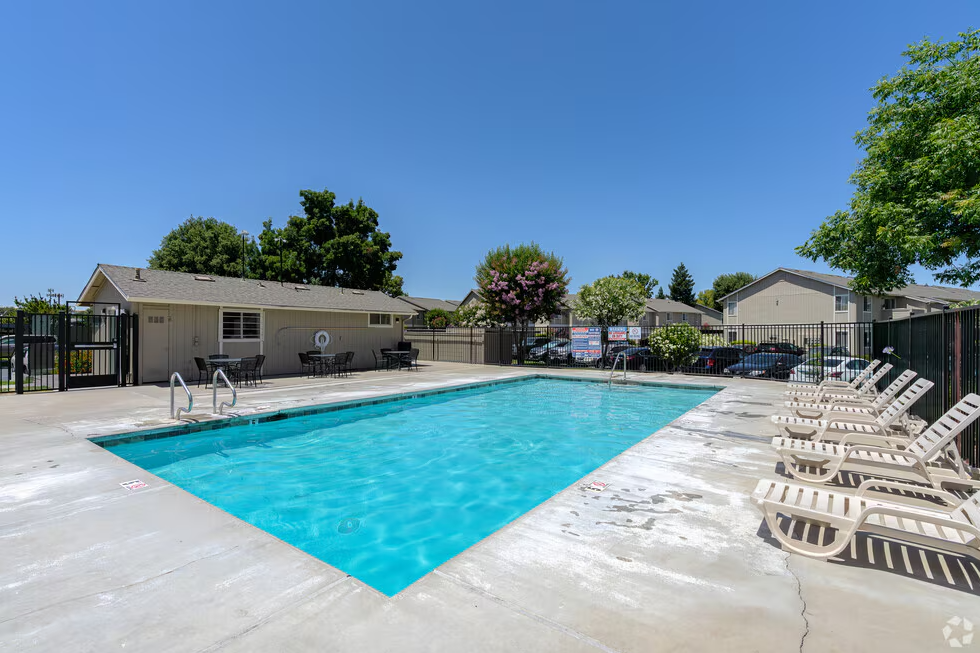 Swimming pool surrounded by concrete, lounge chairs, and small building under a bright blue sky.