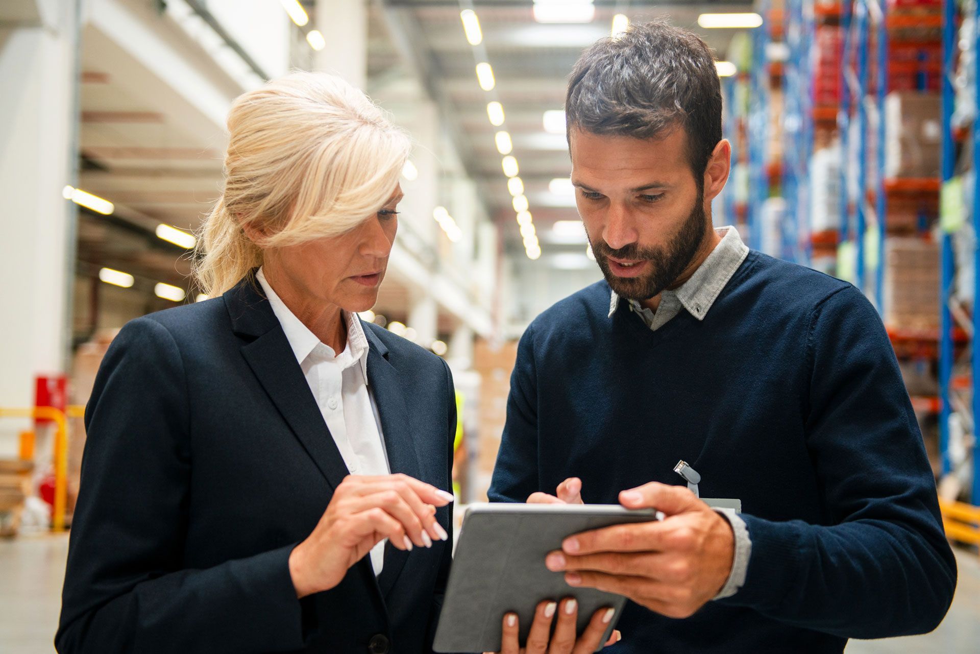 Two people looking at tablet in warehouse. Woman in blazer points, man in sweater looks on.