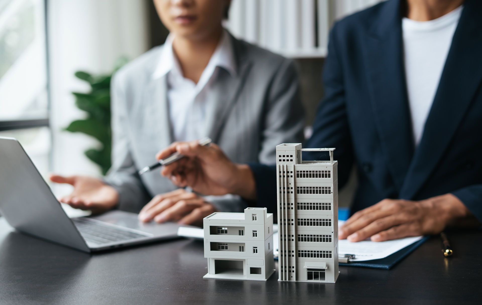 Two people in suits looking at a laptop and small building models on a desk.