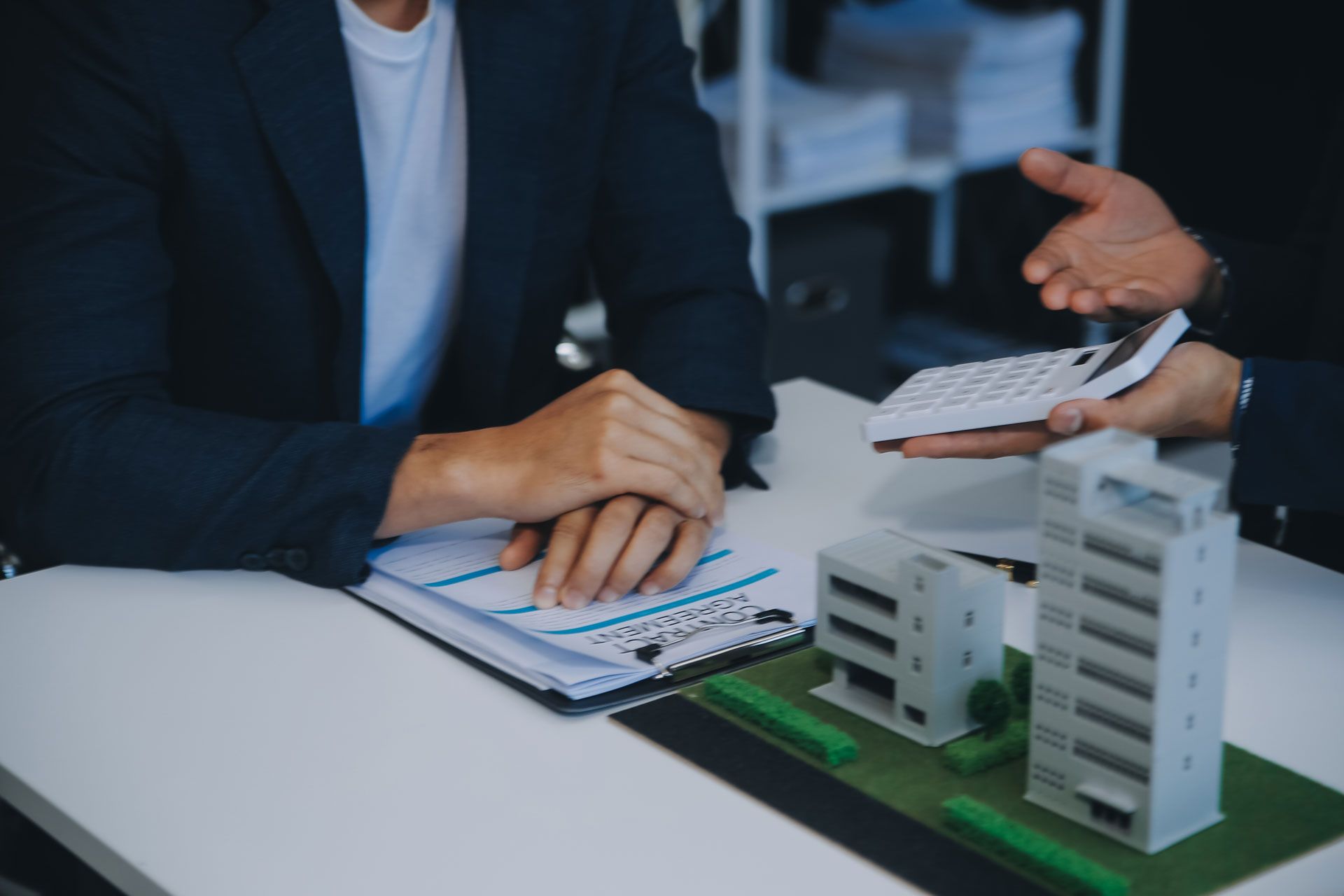 Two people at a desk: one with hands clasped, the other using a calculator, with building models and paperwork.