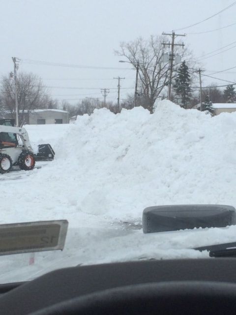 Snow piled high in parking lot with a Bobcat loader; overcast sky and utility poles in background.