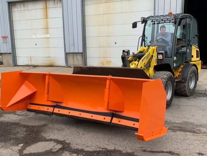 A yellow and black front-end loader with an orange snowplow. A man is driving. The loader is in front of a closed garage.