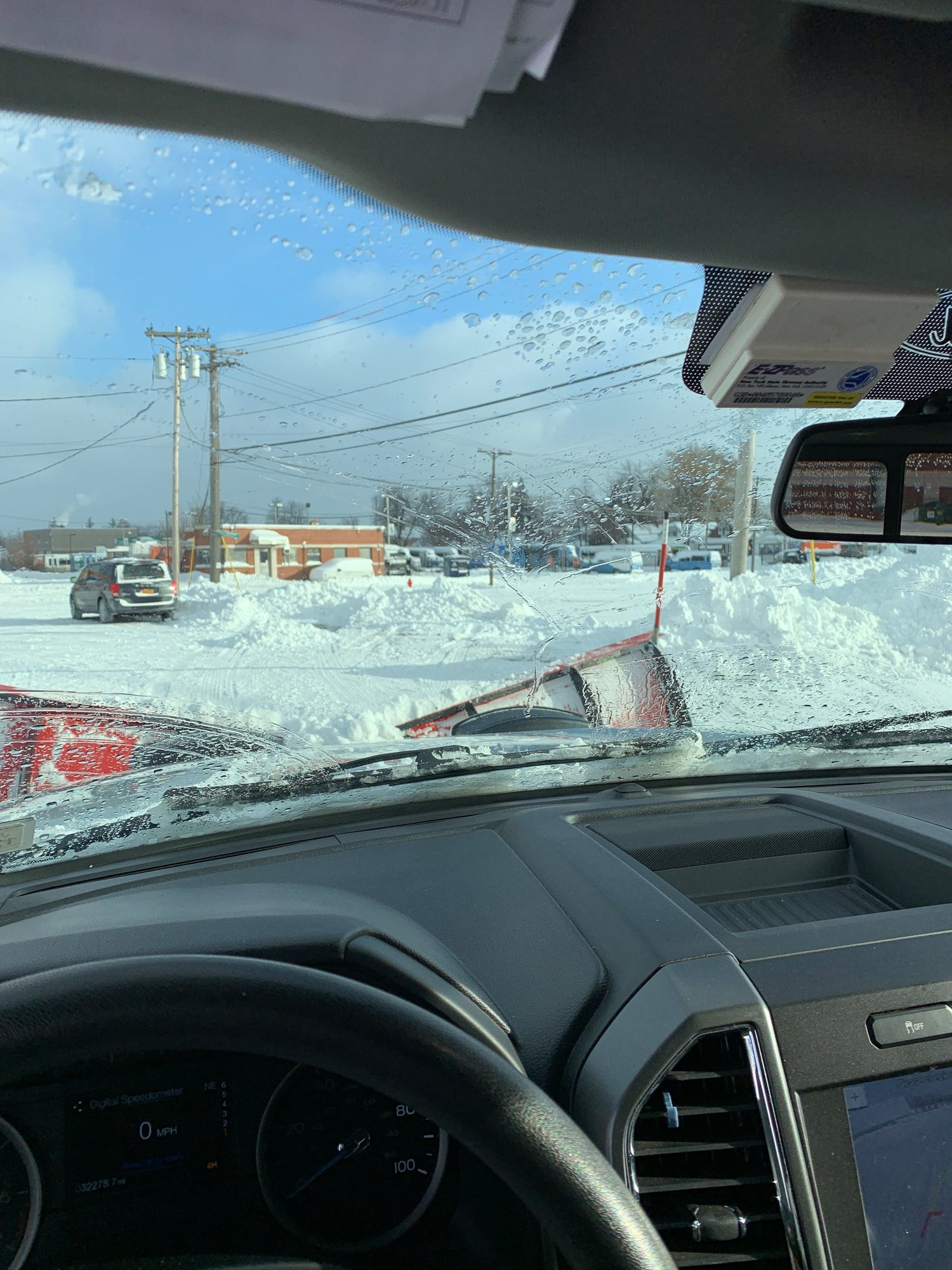 View from inside a truck, watching a snowplow clear a snowy parking lot on a sunny day.