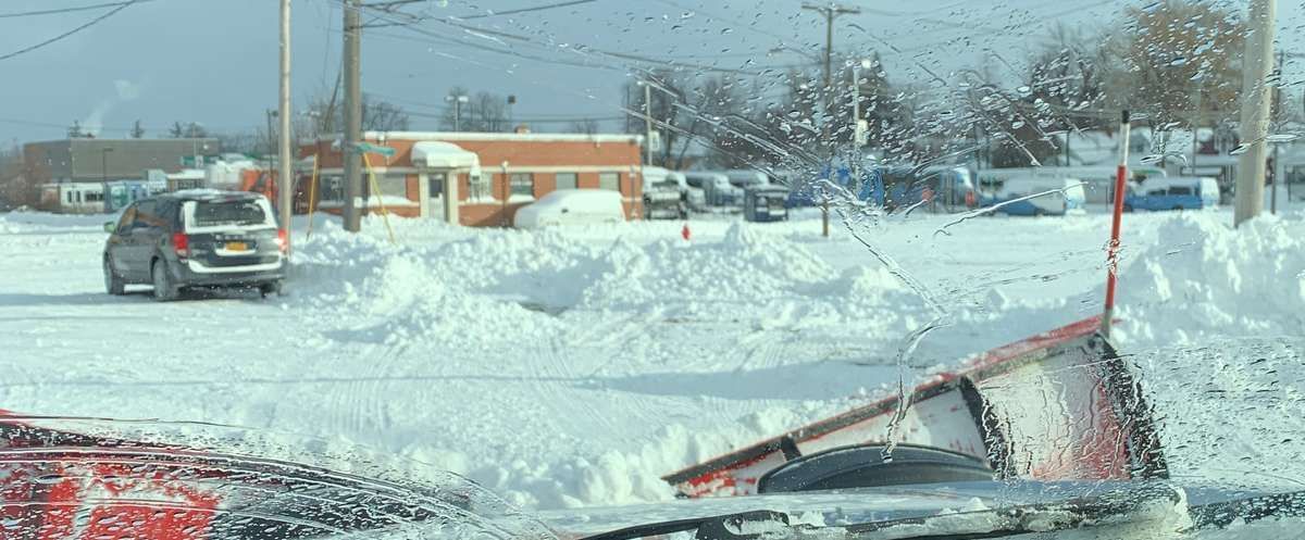 A snowplow clears a street after a blizzard. Buildings and a car are visible in the distance.