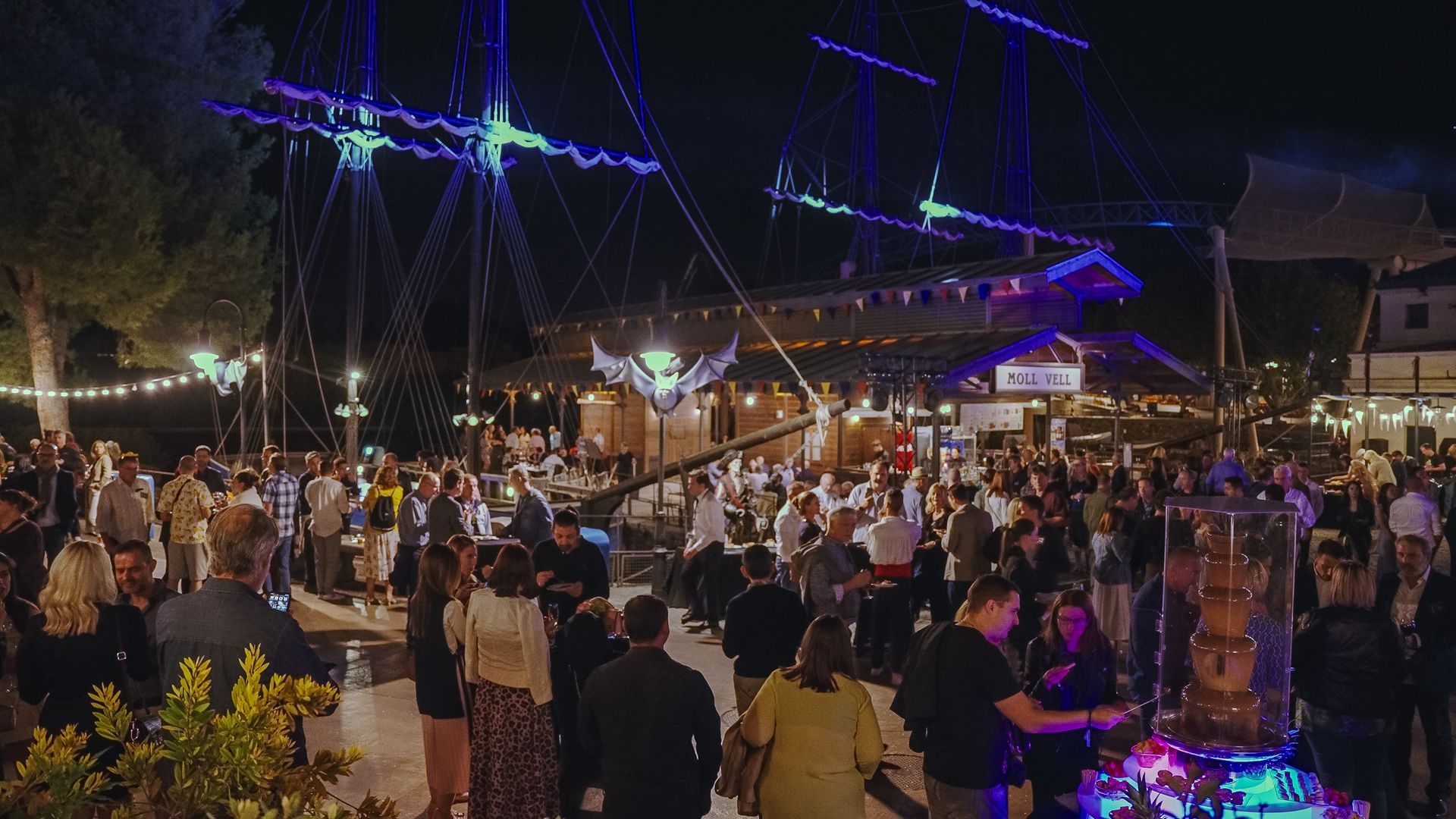 Large outdoor night gathering with a ship-themed structure lit up with colorful lights, and people socializing.