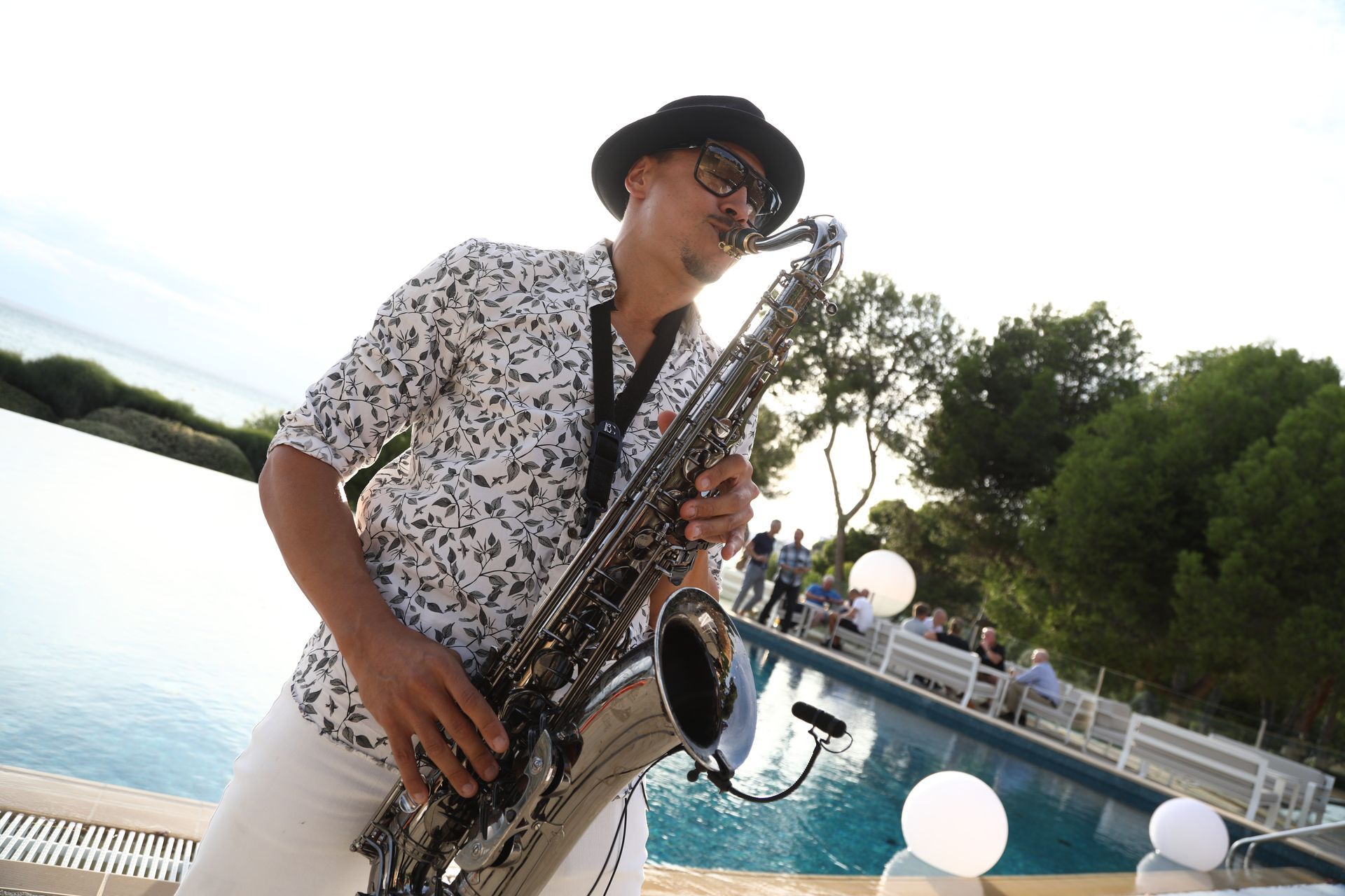Man in sunglasses playing saxophone by pool at outdoor event.