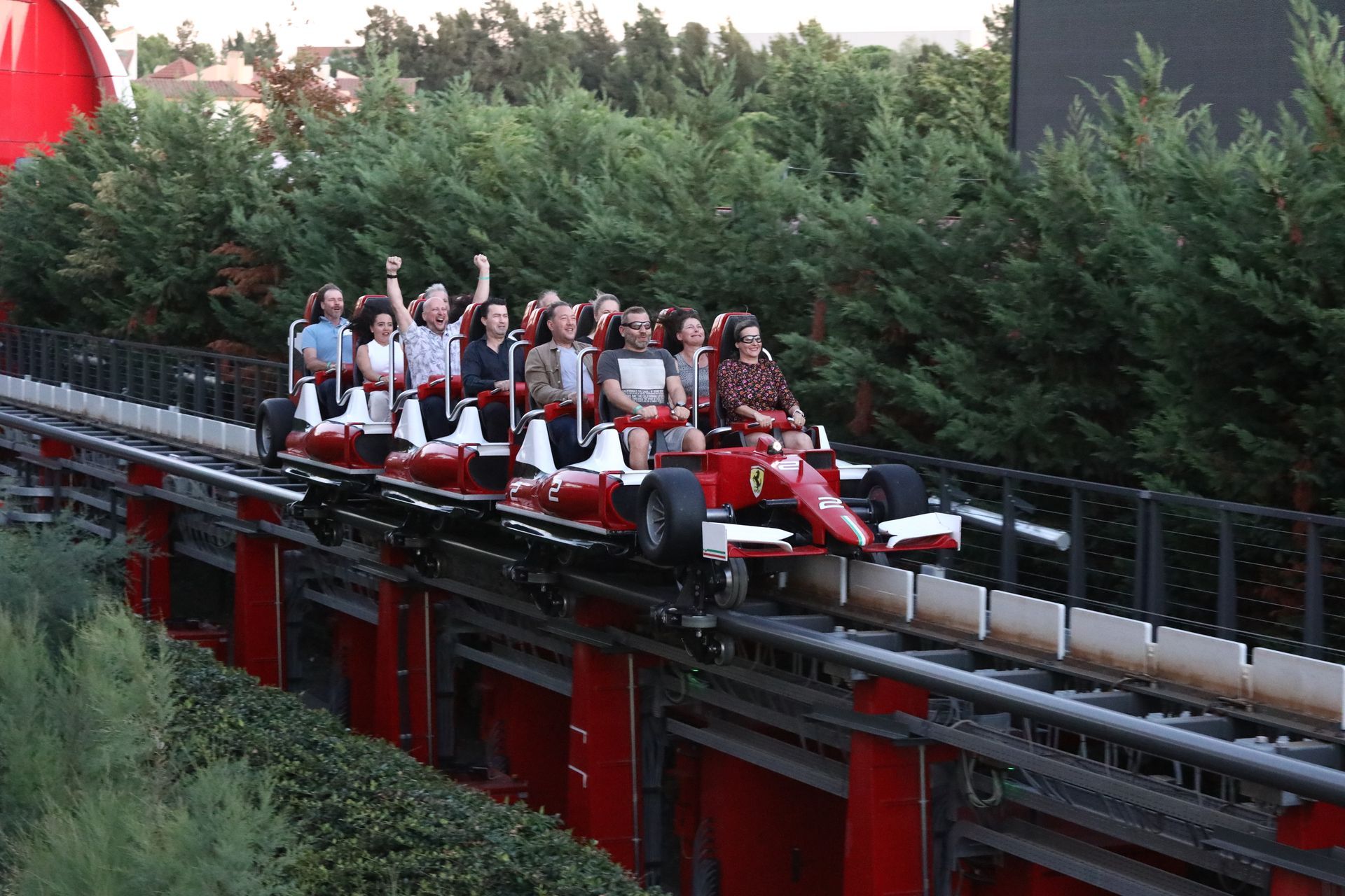 Roller coaster resembling a red Ferrari race car carrying excited riders through a green, outdoor track.