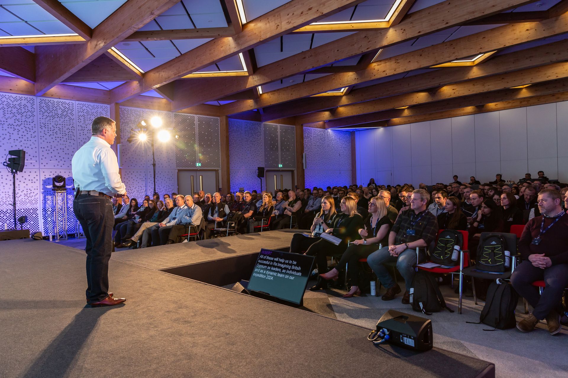 Man speaking on stage to a large audience in a conference hall, with wood beams and blue lighting.