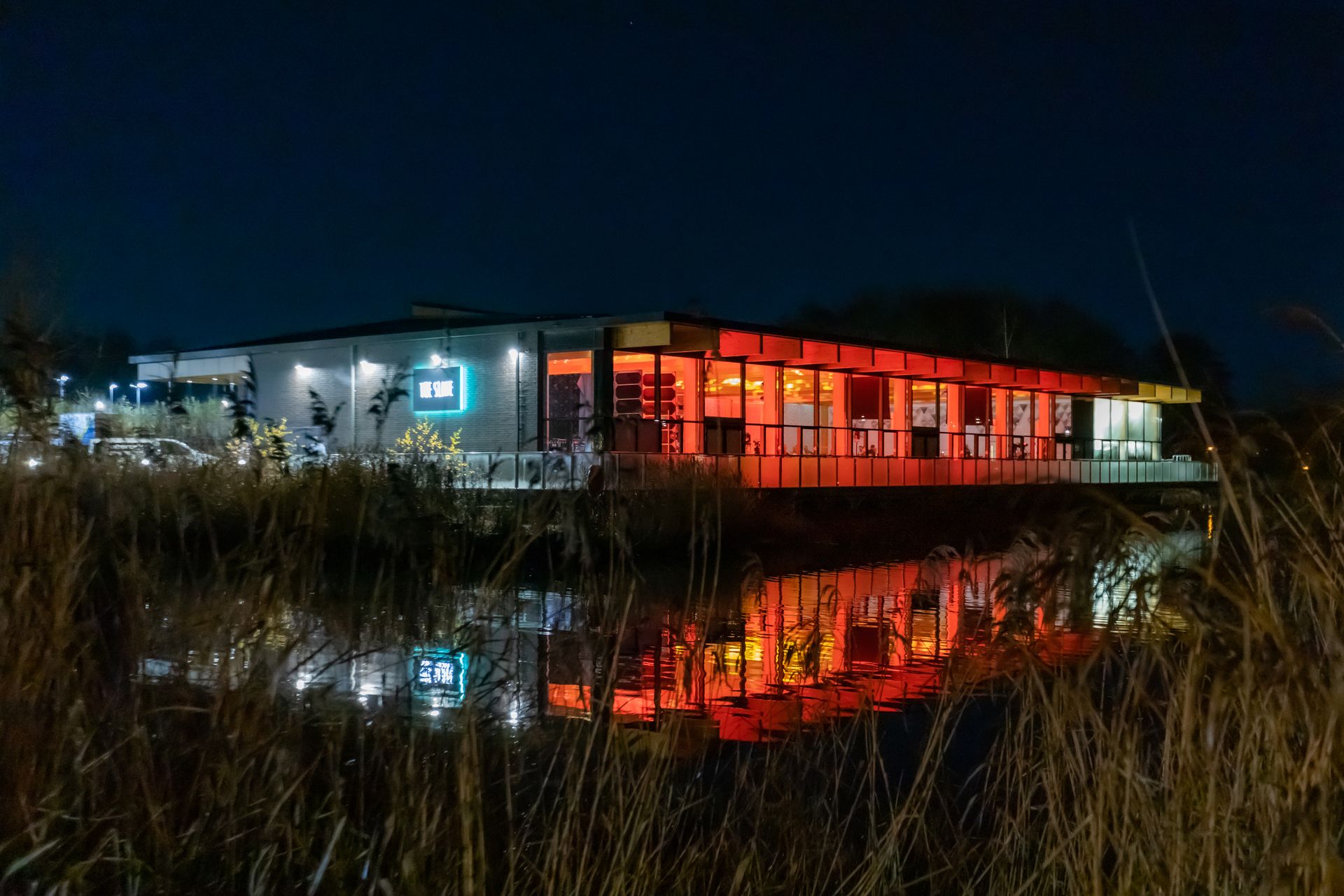 A modern building illuminated with red and blue lights reflects in a dark water at night.