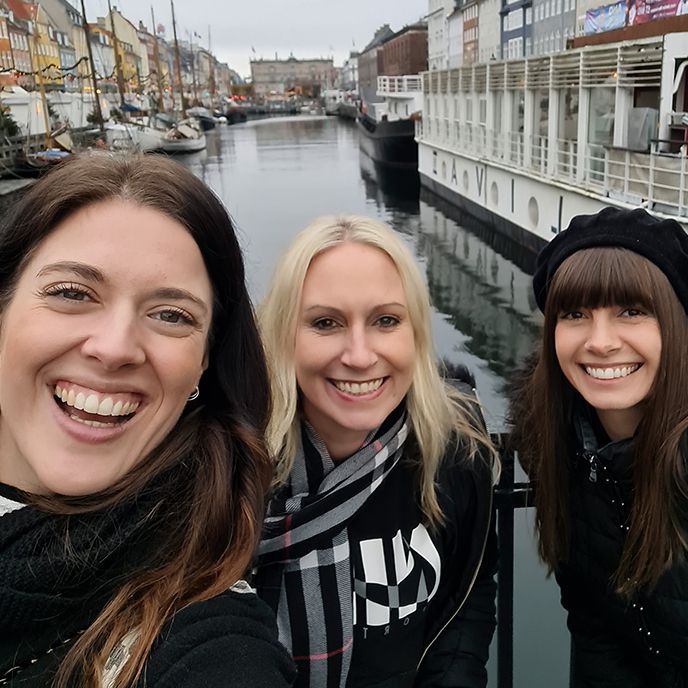 Three women are smiling in front of a body of water