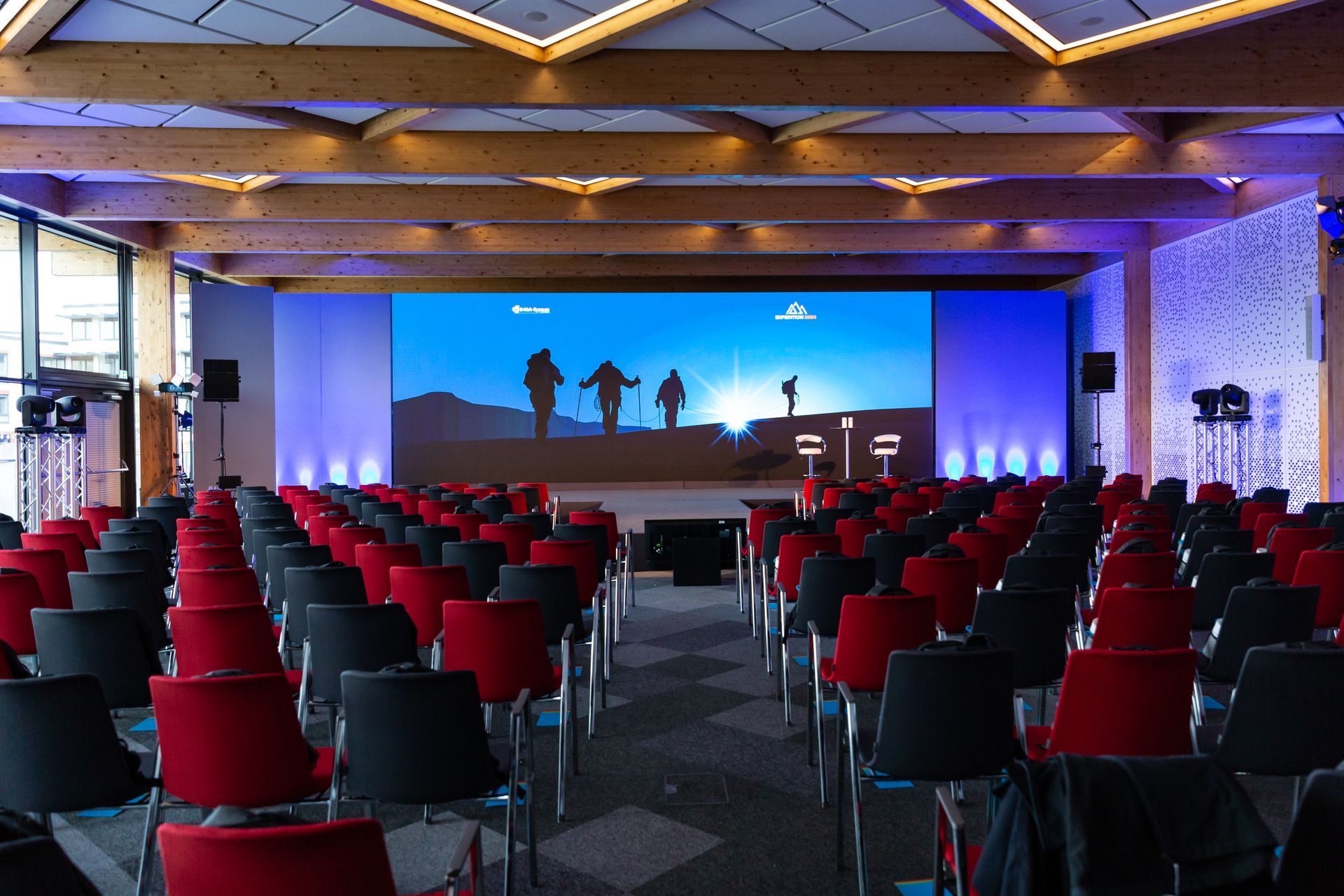 Conference hall with red and black chairs facing a large screen, showing silhouettes on a mountain.