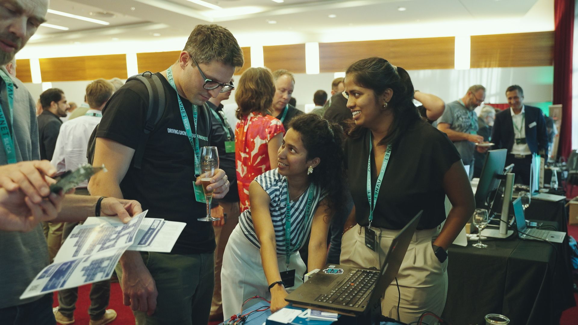 People at an event, looking at a laptop and printed materials. Some have lanyards, indoors.