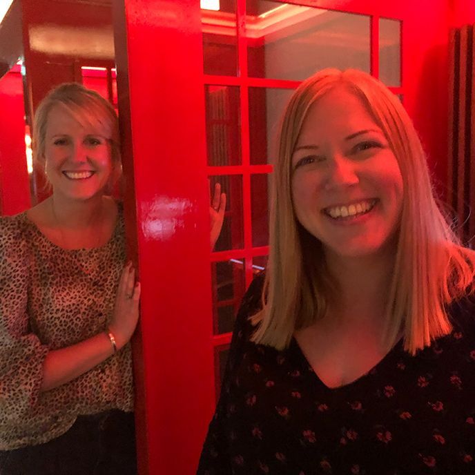 Two women are standing next to each other in front of a red phone booth.