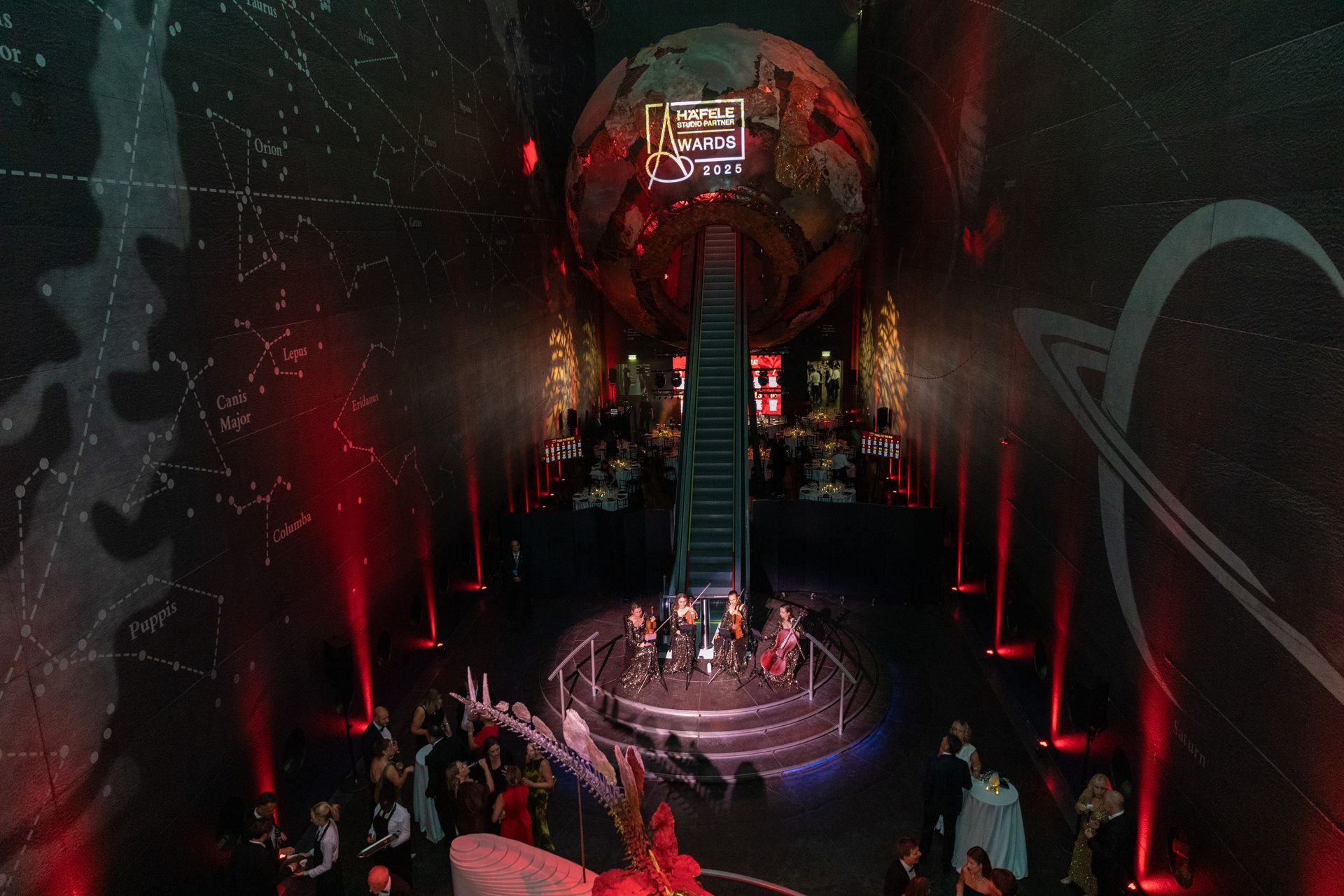 A group of people standing in a futuristic space with a central stage, red lighting, and a large globe.