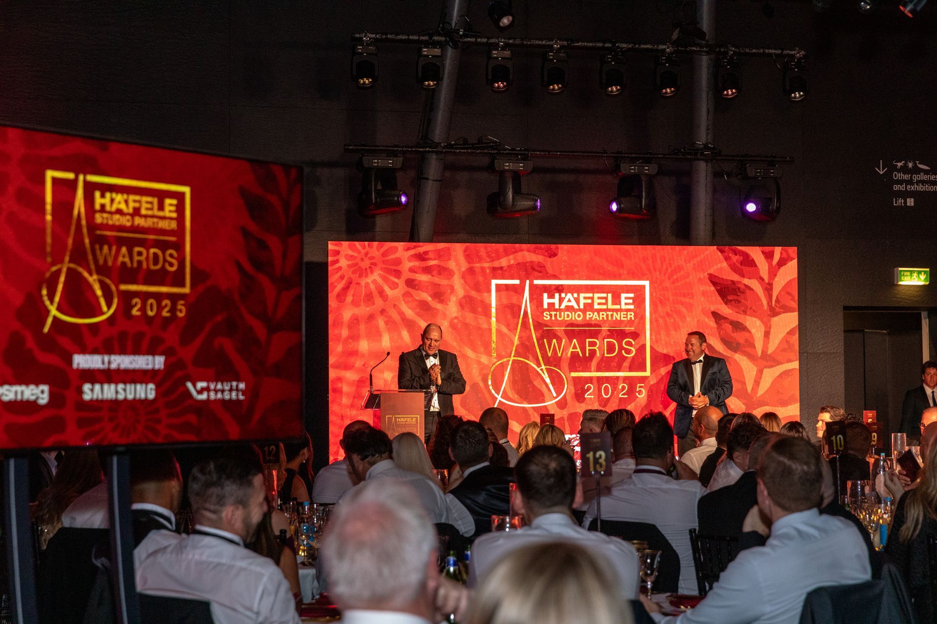 People at the Häfele awards ceremony on a stage with two men at the podium.