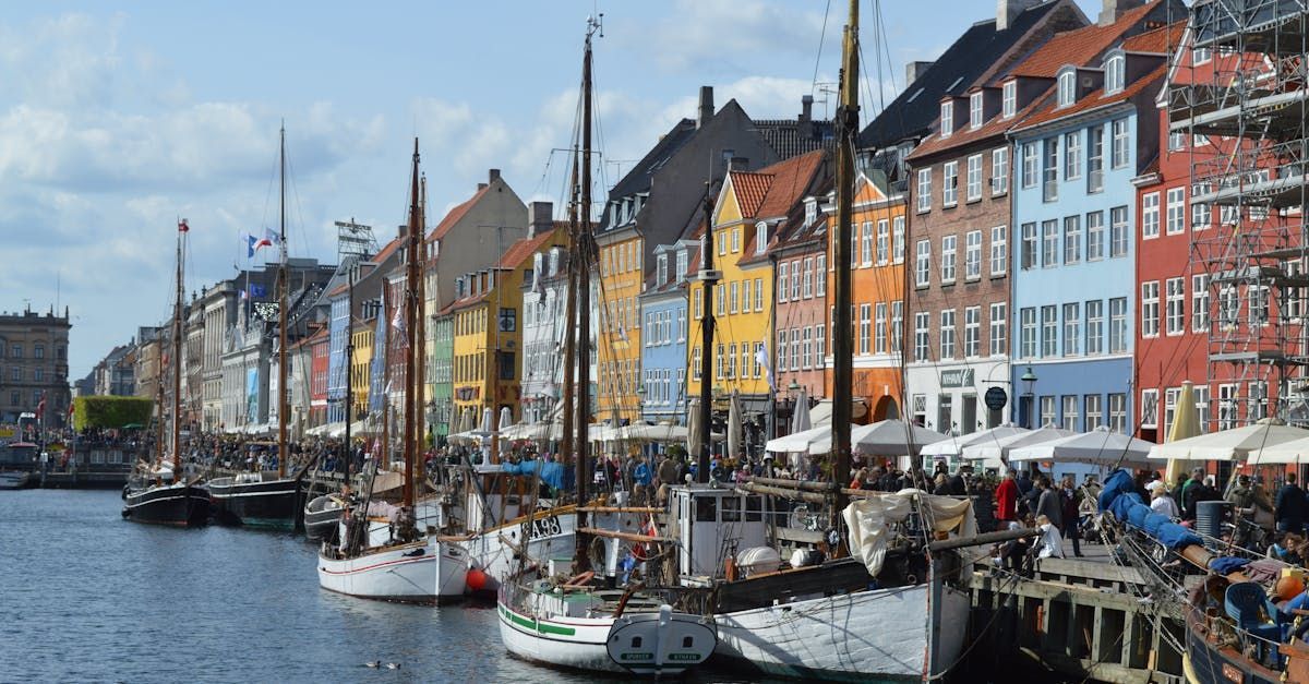 A row of boats are docked in a harbor with buildings in the background.