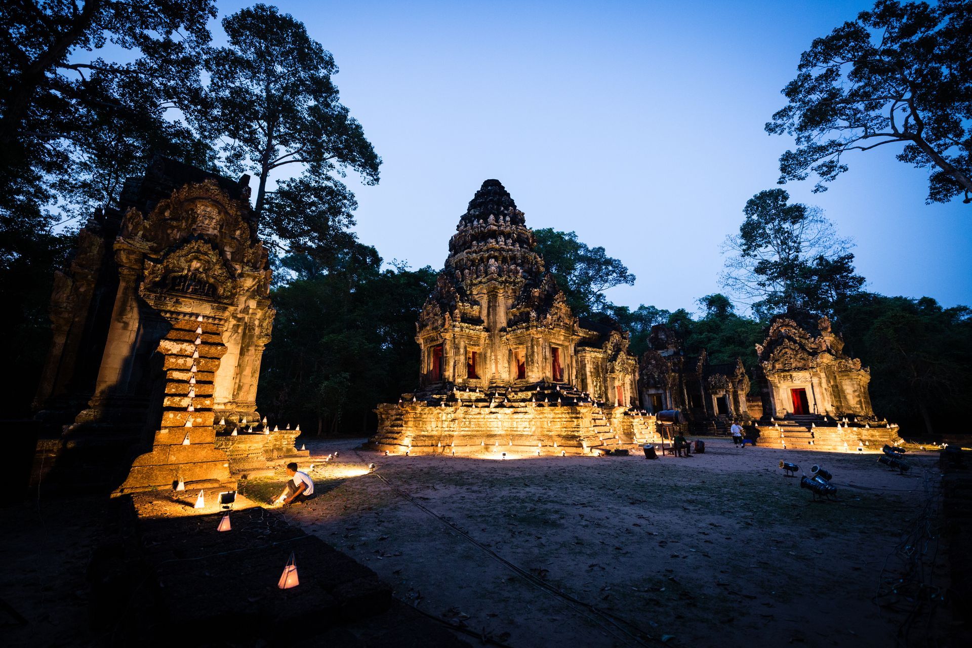 Lit-up ancient temple complex at dusk in Cambodia, surrounded by trees, with warm lighting casting shadows.
