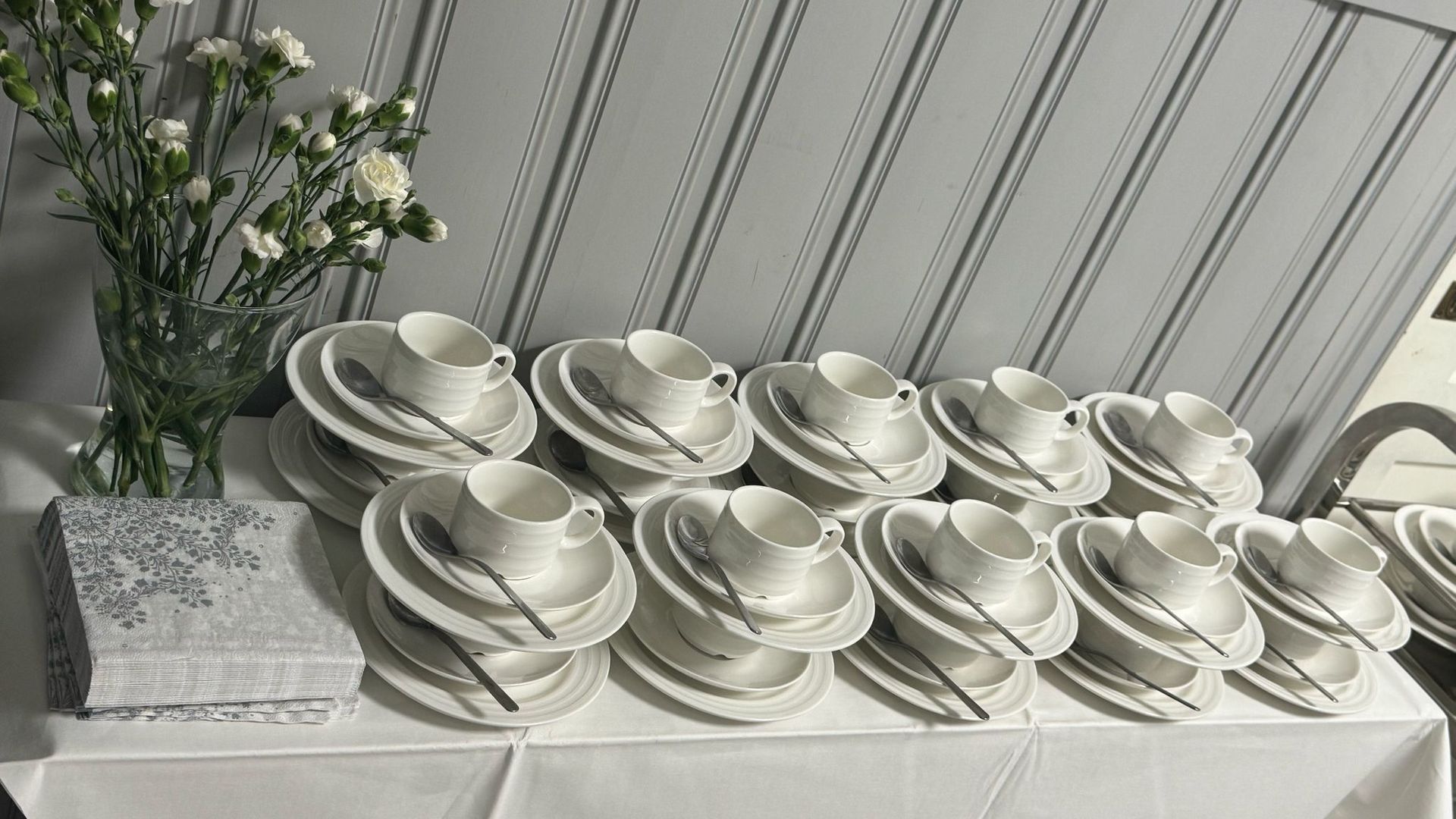 Pöytä, johon kuuluu valkoisia teekuppeja, lautasia, lusikoita ja lautasia; maljakko valkoisia kukkia ja koristeellinen rasia.