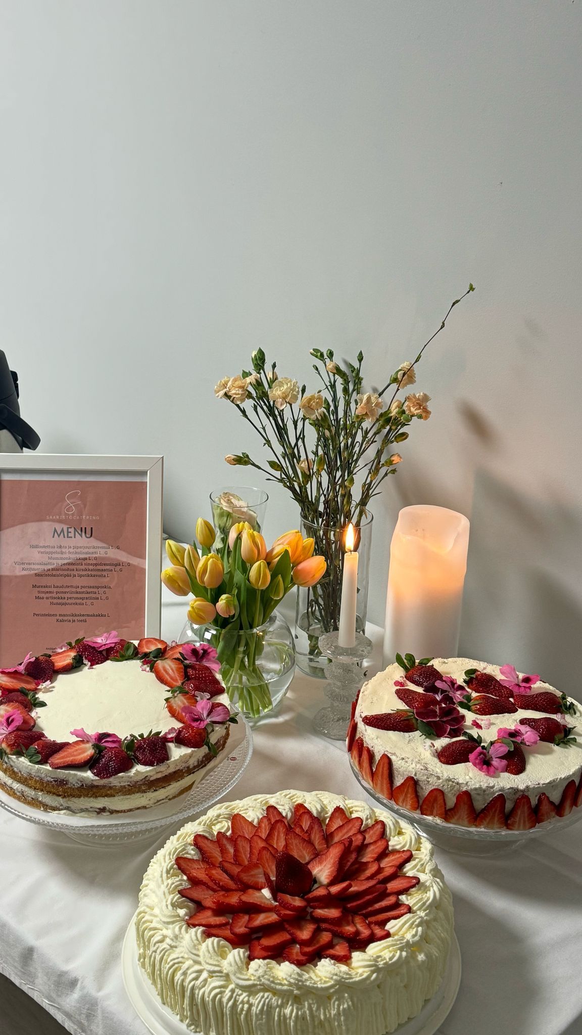 Kolme koristeltua kakkua kukilla, kehystetty todistus, kukkia, kynttilöitä pöydällä, jolla on valkoinen pöytäliina.