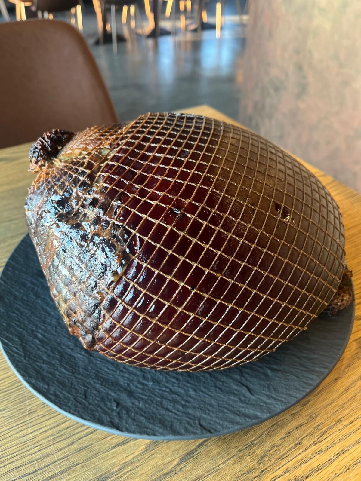 Suuri, pyöreä, kypsennetty liha, käärittynä ruskeaan verkkoon, on tummalla lautasella vaalealla puupöydällä.