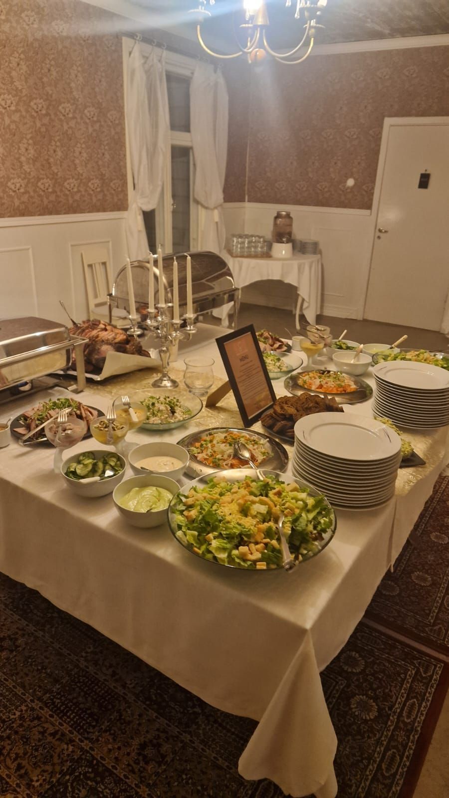 Buffetpöytä erilaisilla annoksilla huoneessa, jossa on kattokruunu, valkoinen pöytäliina ja beigenväriset seinät.