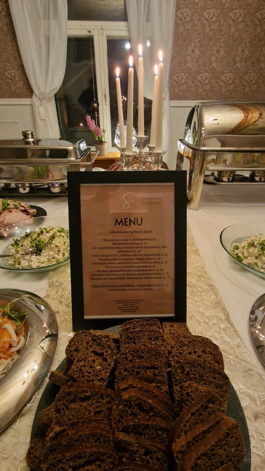 Buffetpöytä, jossa on ruokalista, leipää, salaatteja ja kuumailmaruokia kynttilöiden alla.