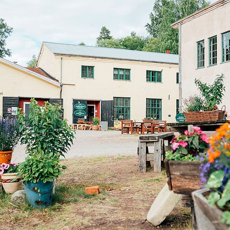 Sisäpiha, jossa on rakennuksia, kasveja, pöytiä ja puisia yksityiskohtia. Keltaiset seinät, vihreät listat ja pilvinen taivas.