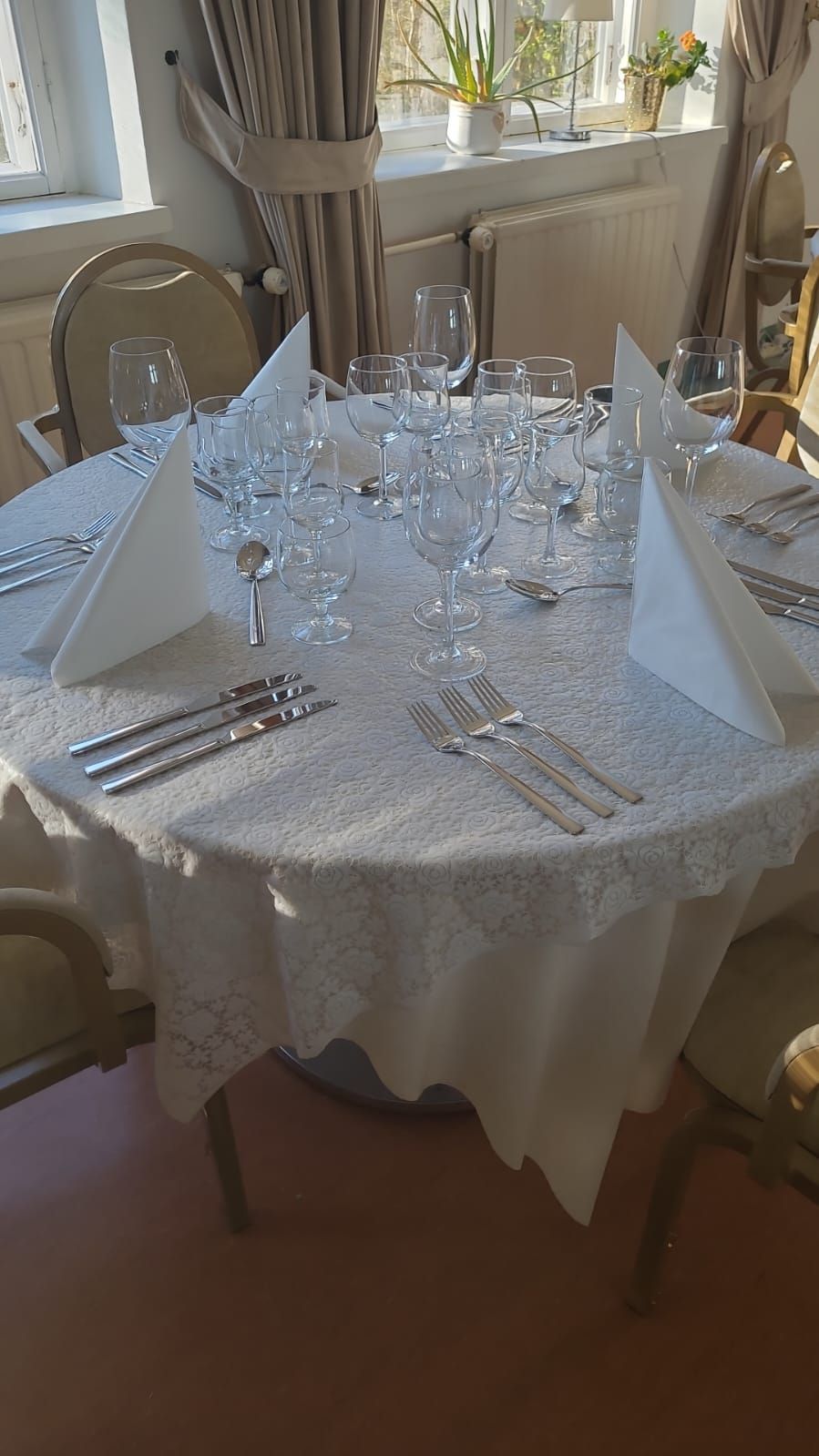 Pyöreä pöytä katettu aterialle, jossa on valkoinen pitsipöytäliina, aterimet, lasit ja taitetut lautasliinat.