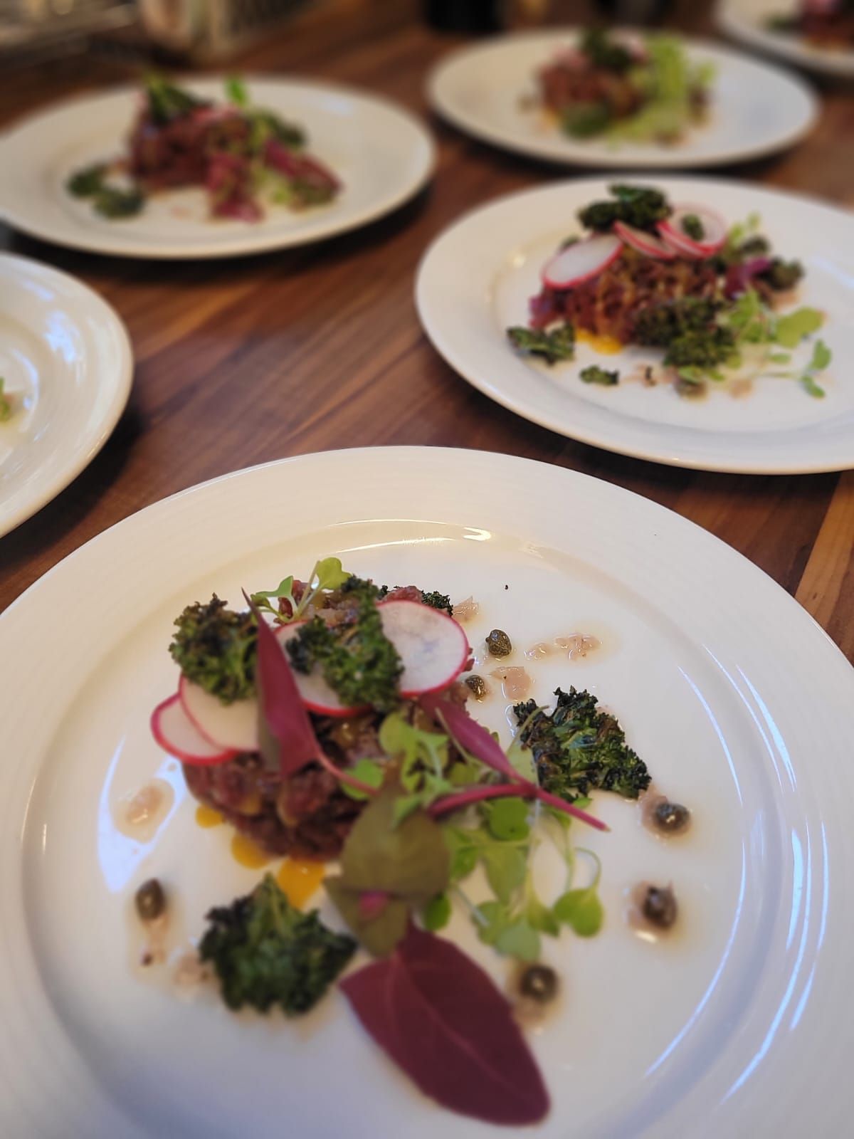 Useita lautasia värikkäitä ruokia puupöydällä, mahdollisesti ravintola. Jokaisella lautasella on liharuoka, vihanneksia ja lisukkeita.