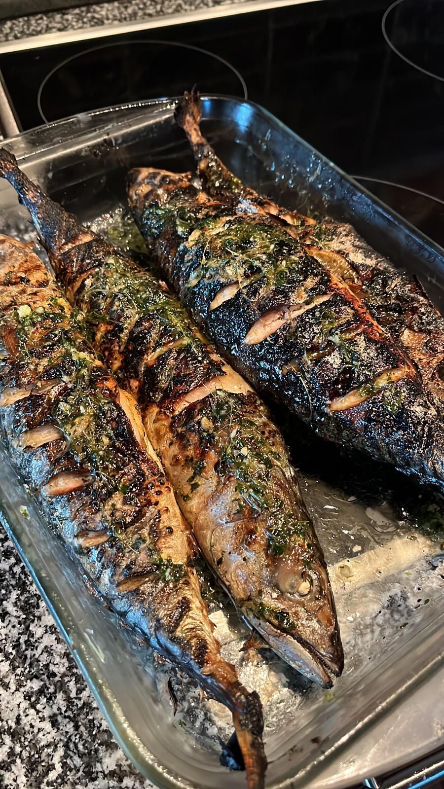 Grillattu kokonaisena kala lasisessa uunivuoassa, yrttikoristeena ja grillimerkeillä.