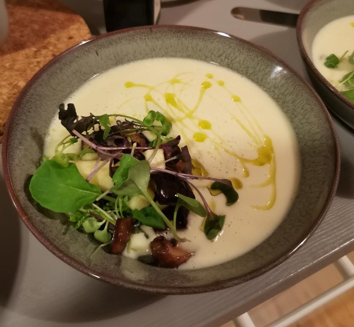 Kermainen valkoinen keitto harmaassa kulhossa, koristeltuna vihreillä, sienillä ja oliiviöljyllä.