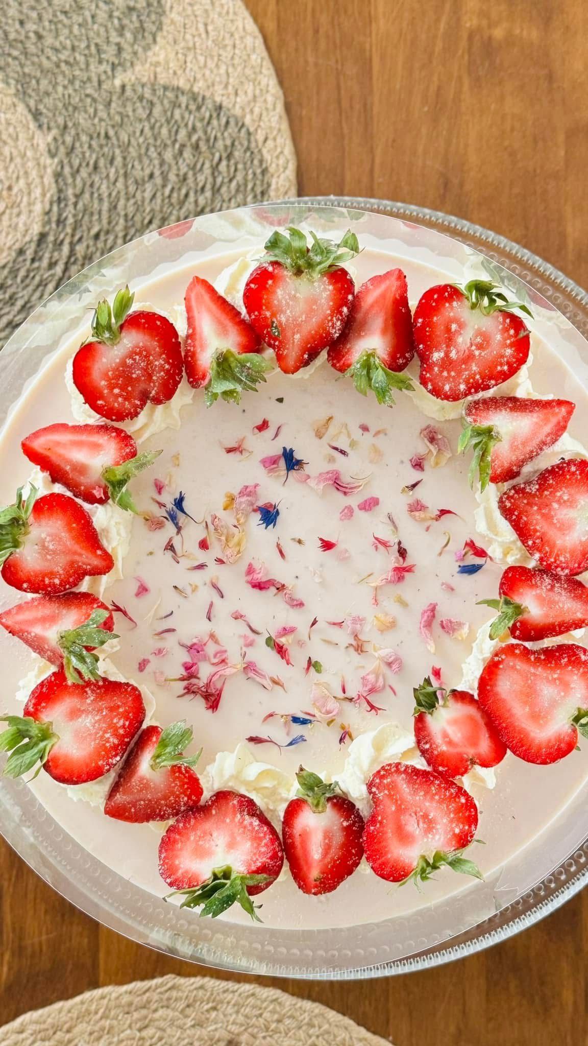 Mansikkakakku kermavaahdolla, ruusun terälehdillä ja viipaloiduilla mansikoilla päällä puupöydällä.