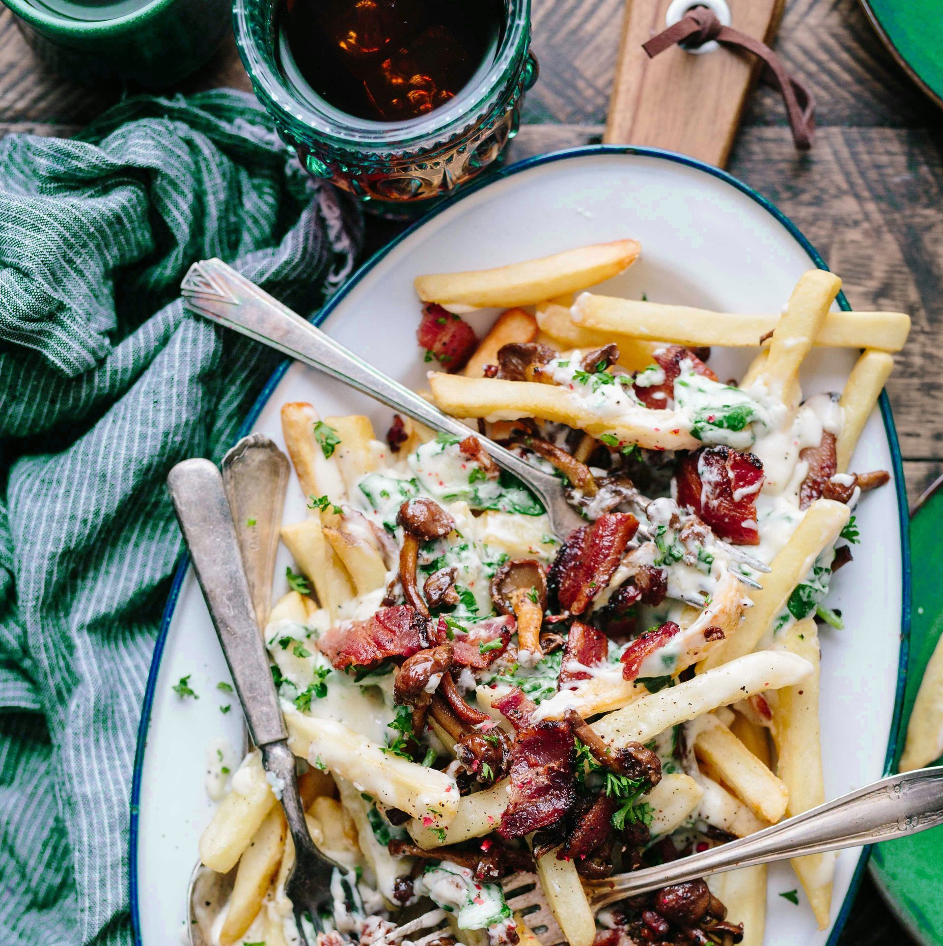 Ranskalaiset perunat pekonilla, juustokastikkeella ja persiljalla valkoisella tarjoiluvadilla, jossa on vihreitä yksityiskohtia ja aterimia.